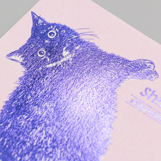 A close-up of a printed illustration of a black cat on a pale pink paper background, with a textured metallic purple foil stamp effect over the cat's fur.