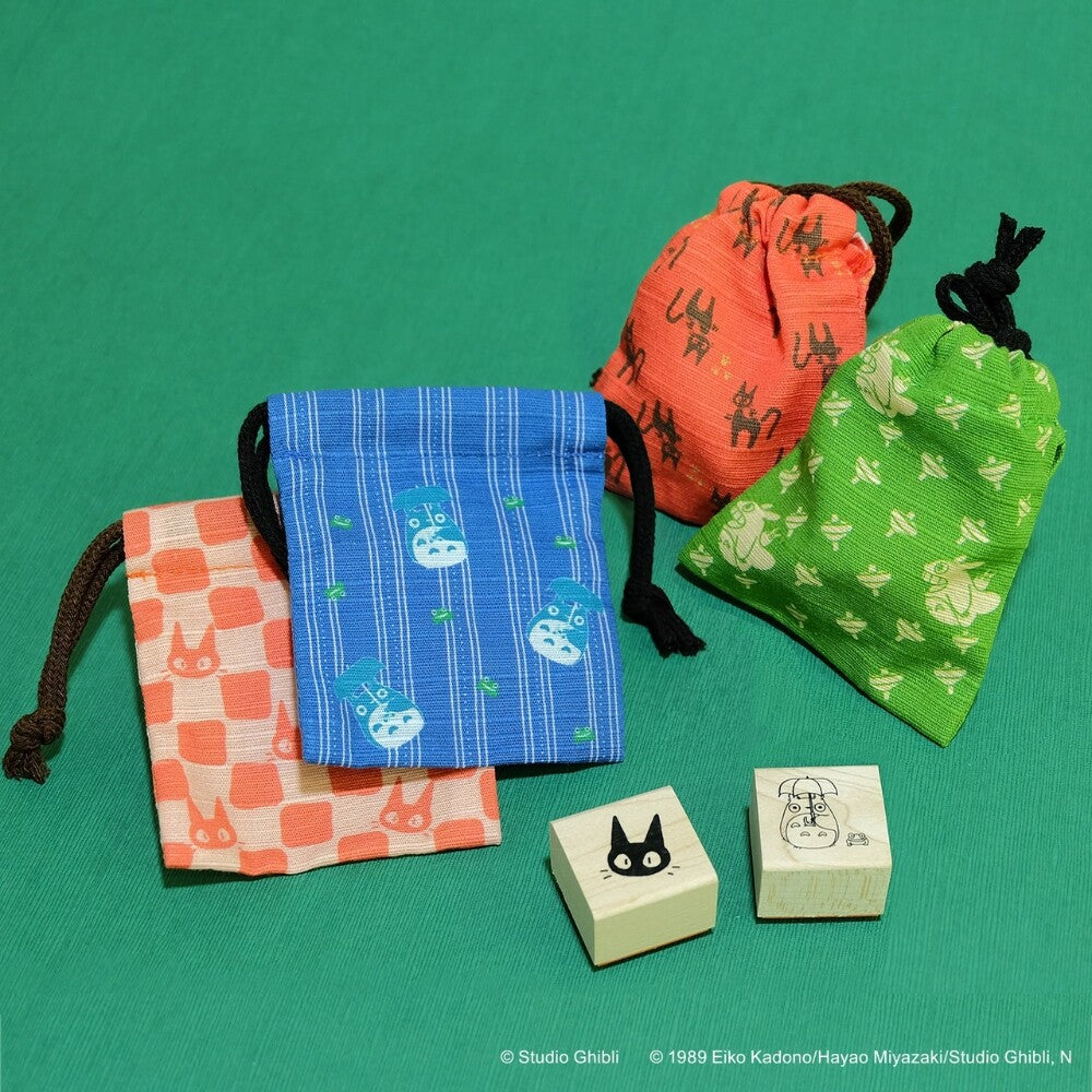 Four fabric drawstring pouches in orange, blue, green, and peach with cartoon prints, alongside two small wooden stamps, arranged on a green background. Studio Ghibli copyright text appears at the bottom.