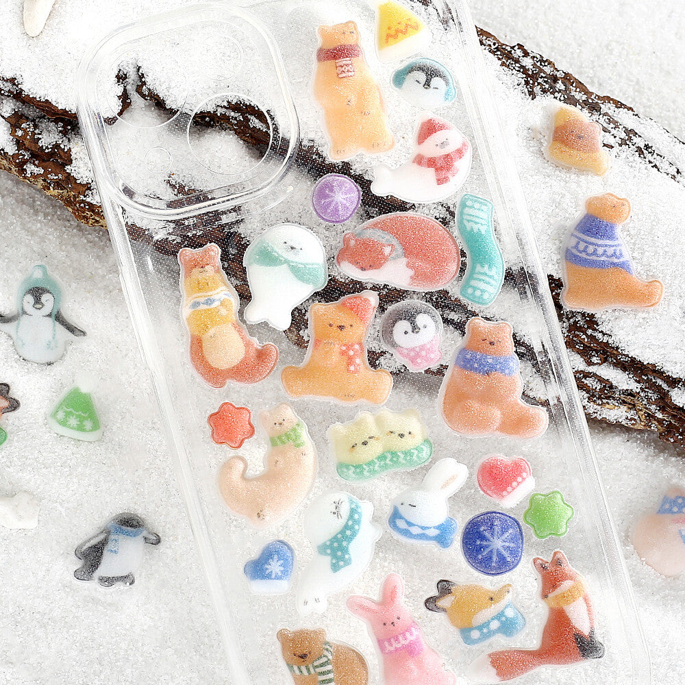 A clear phone case decorated with colorful winter-themed cartoon stickers, including penguins, bears, rabbits, and snowflakes, displayed on a snowy surface with a log in the background.