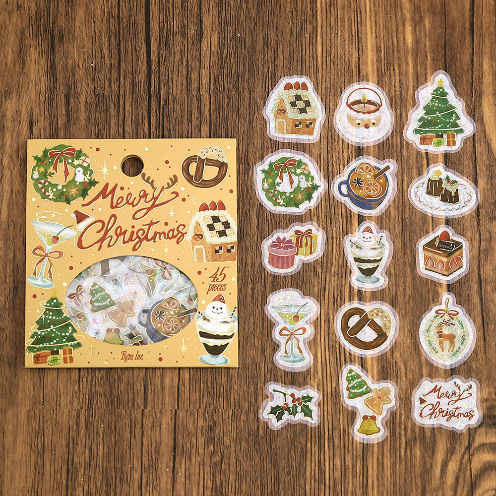 A Christmas-themed sticker pack in a decorative yellow sleeve with illustrations of ornaments, a gingerbread house, a martini glass, and the words 'Merry Christmas.' A circular window reveals assorted stickers inside.