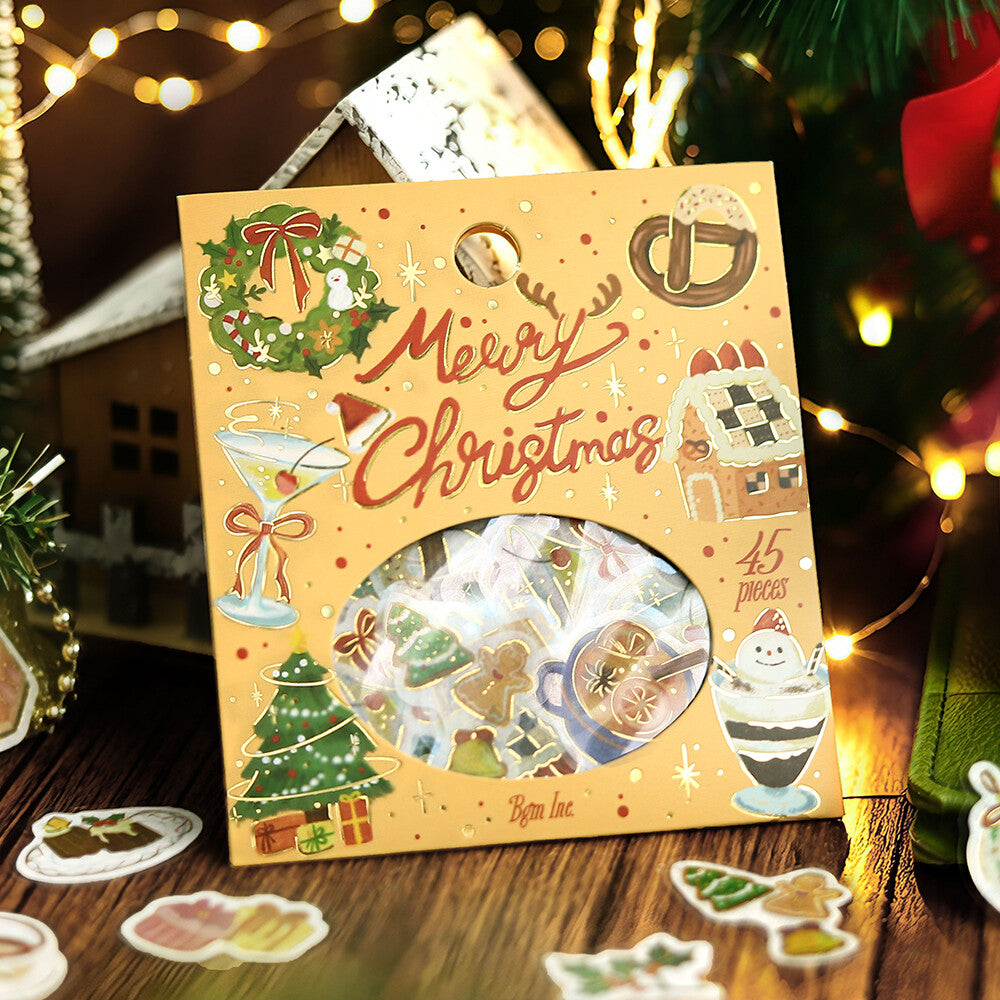 A Christmas-themed sticker pack in a decorative yellow sleeve with illustrations of ornaments, a gingerbread house, a martini glass, and the words 'Merry Christmas.' A circular window reveals assorted stickers inside.
