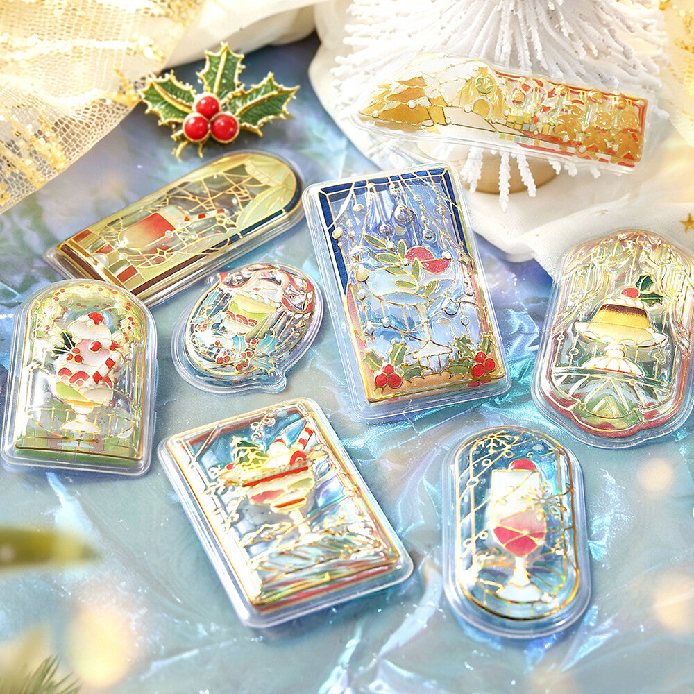 A set of decorative acrylic stickers with illustrated winter and holiday scenes, arranged on a light blue surface with festive props including a holly sprig and soft fabric.