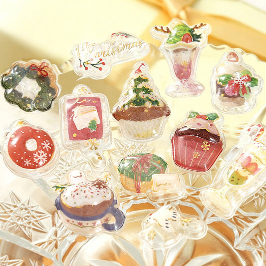 A set of clear acrylic stickers shaped like Christmas treats and desserts, including cupcakes, a donut, a holiday wreath, a milkshake, and a mug with a winter hat, displayed on a crystal tray.