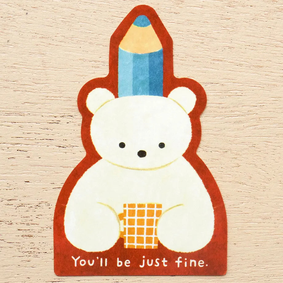 A die-cut sticker featuring a cute white polar bear holding a yellow-orange checkered cup, with a blue pencil on its head and the text 'You'll be just fine.' at the bottom, shown on a light wood background.