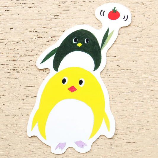 A die-cut sticker featuring two cute cartoon penguins on a light wood background. The foreground penguin is bright yellow with a white belly, black eyes, a small red beak, and light purple feet.