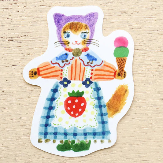 A die-cut sticker featuring a whimsical hand-drawn illustration of a cat dressed in a colorful outfit and holding an ice cream cone, placed on a light wood background.