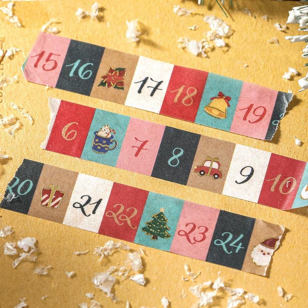 Three colorful paper advent-themed washi tape strips laid diagonally on a textured yellow surface with small white decorative flakes. The strips show numbered sections from 15 to 24 with holiday illustrations including a poinsettia, bell, car, tree, and Santa.