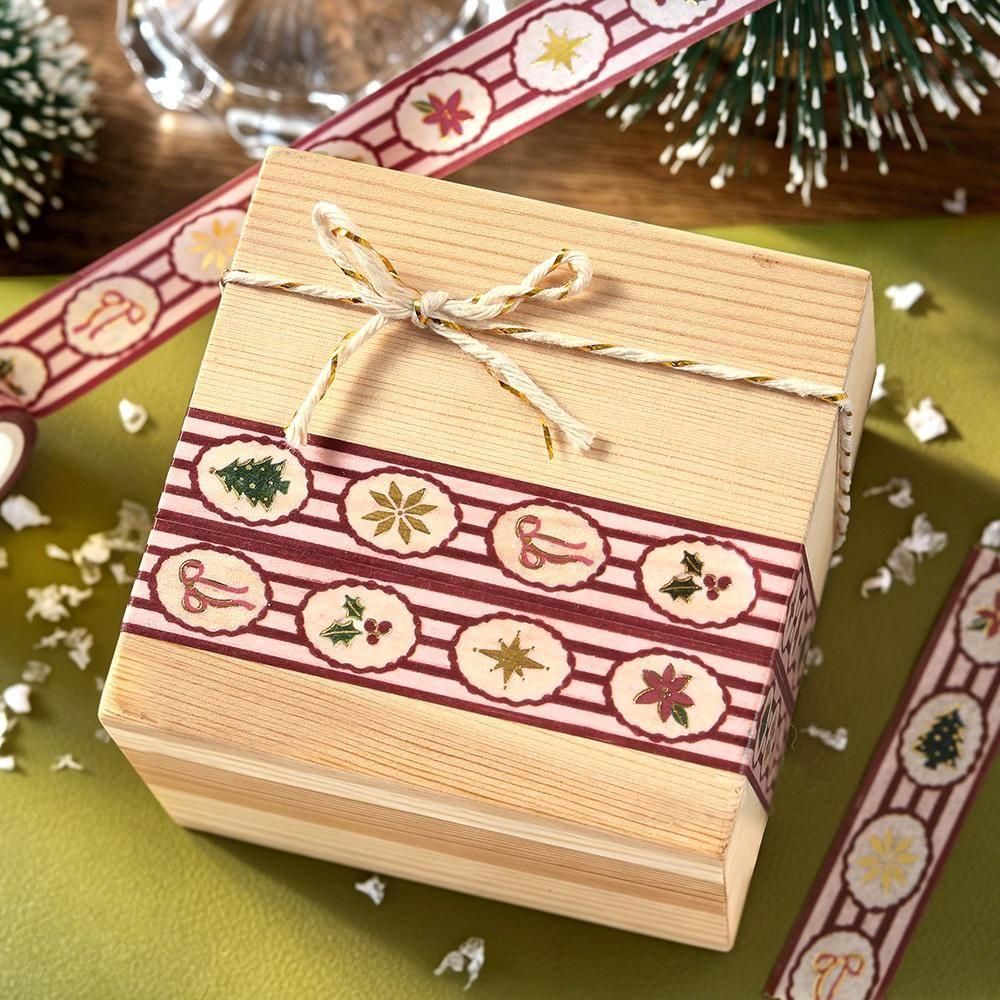 A square wooden gift box with a light natural wood finish, decorated with a red and white holiday patterned washi tape and set on a green surface with festive props.