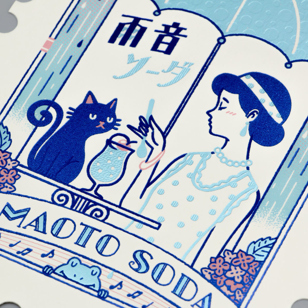 Stylized illustration of a woman, cat, and umbrella with 'AMADO SODA' text on a foil-stamped postcard.