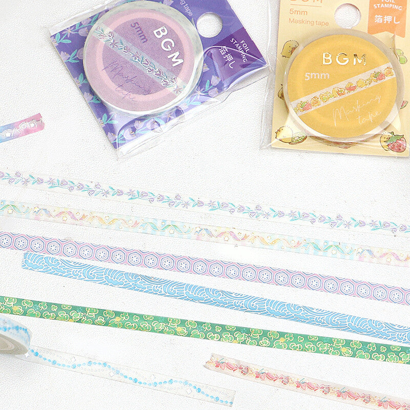 Several rolls of decorative washi tape are laid out on a white surface, showing pastel floral and geometric patterns in blue, green, pink, purple, and yellow. Two packaged rolls are visible at the top, labeled BGM 5mm.