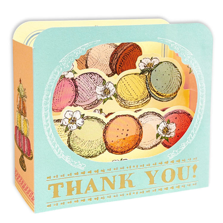 A square greeting card box with a pastel blue front, decorated with an illustration of colorful macarons, white flowers, and the words 'THANK YOU!' in gold. The side panel is peach-orange with a small macaron illustration.