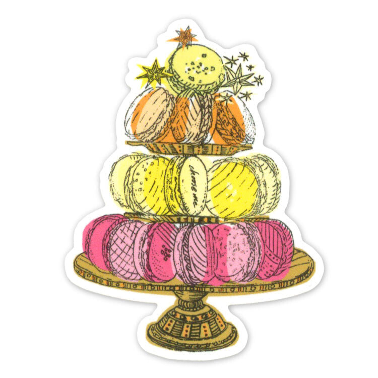 A die-cut sticker featuring an illustration of three tiers of macarons on a decorative stand, with small stars and leaves around the top. The macarons are pink, yellow, and orange, with gold outlines.