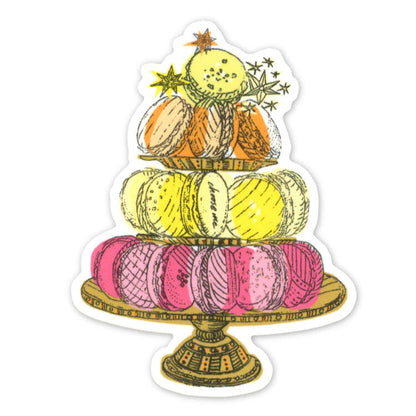 A die-cut sticker featuring an illustration of three tiers of macarons on a decorative stand, with small stars and leaves around the top. The macarons are pink, yellow, and orange, with gold outlines.
