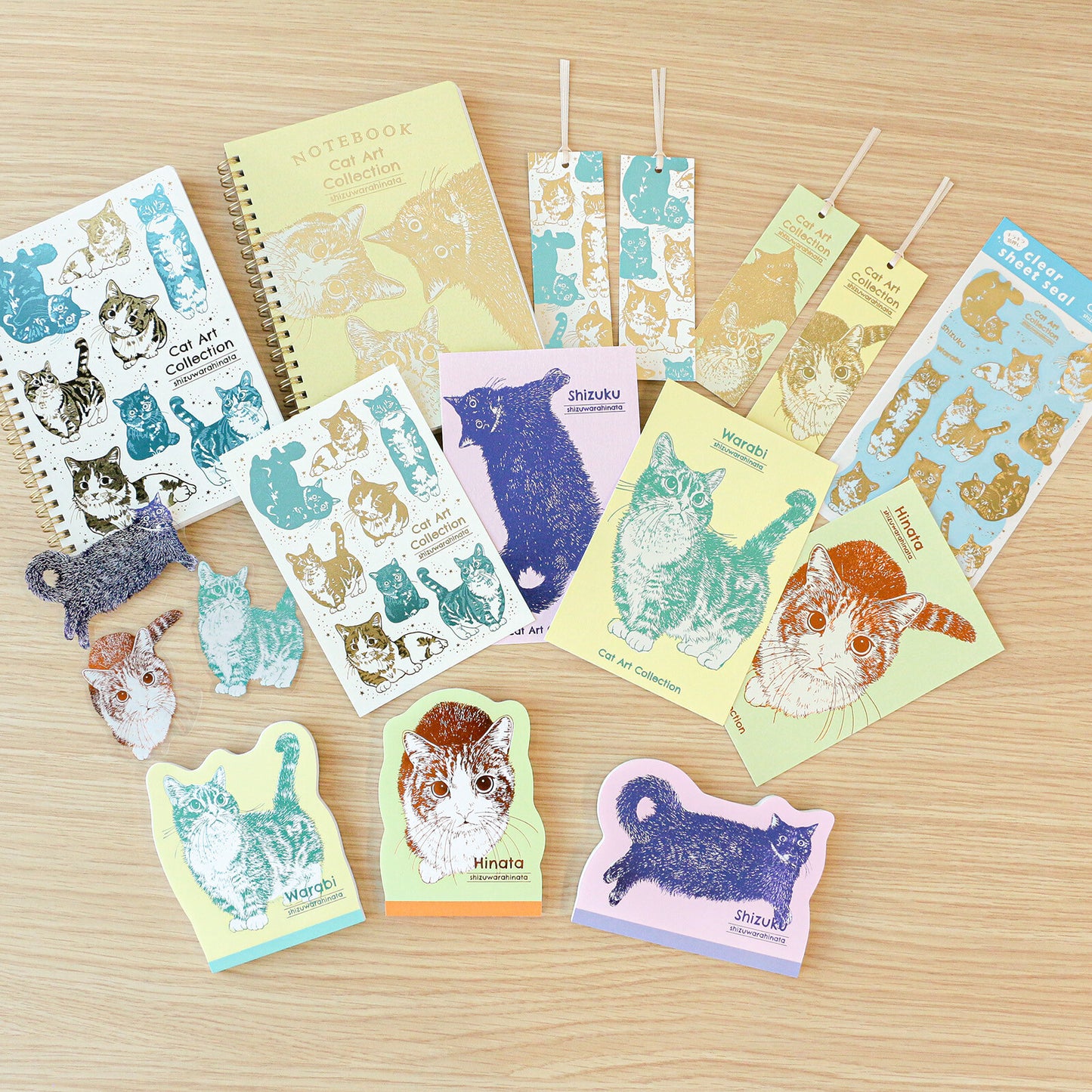 A flat-lay collection of cute cat-themed stationery items on a light wood background, including a notebook, sticker sheets, bookmarks, cards, and die-cut stickers.