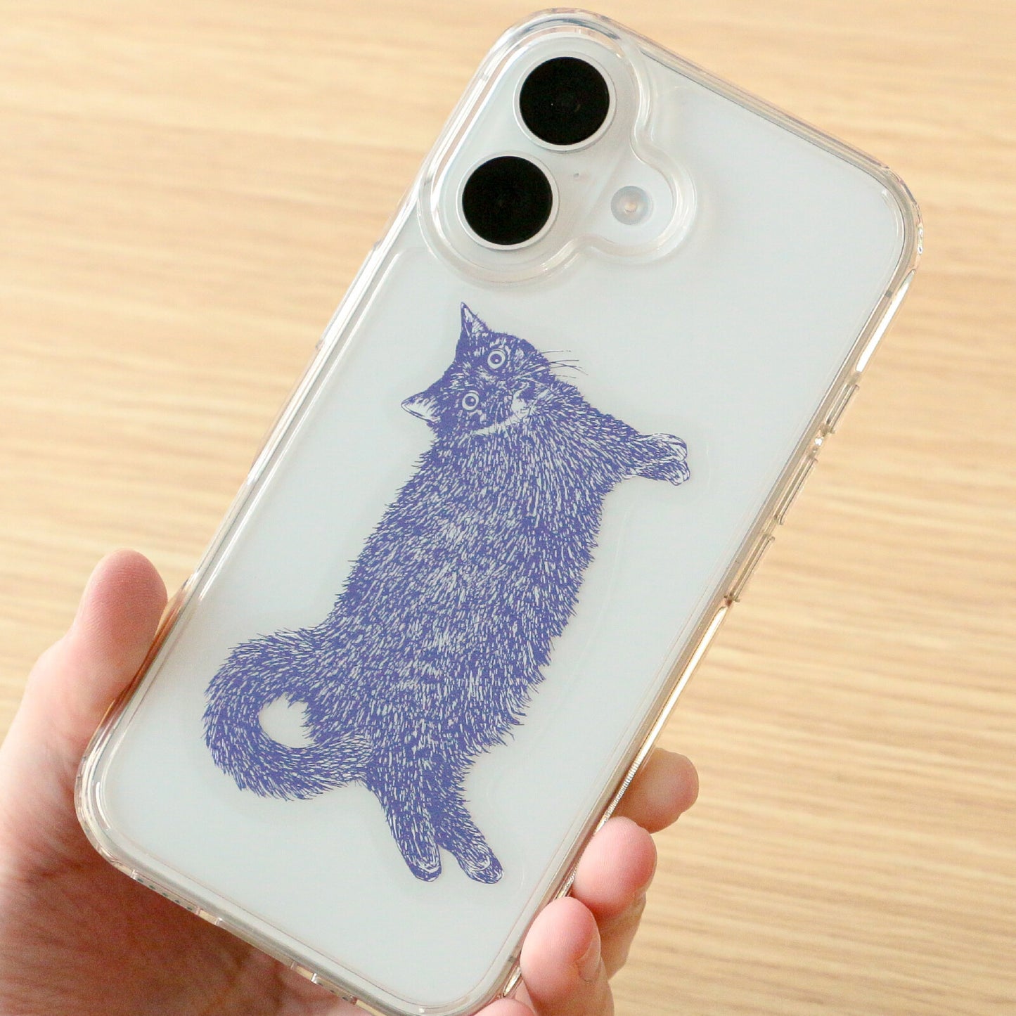 A hand holding a clear phone case with a purple foil line-art illustration of a cat, shown against a light wood background.