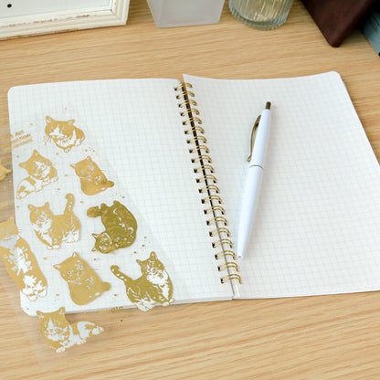 Open notebook with gold cat illustrations on one page and a pen on the other, on a wooden surface.