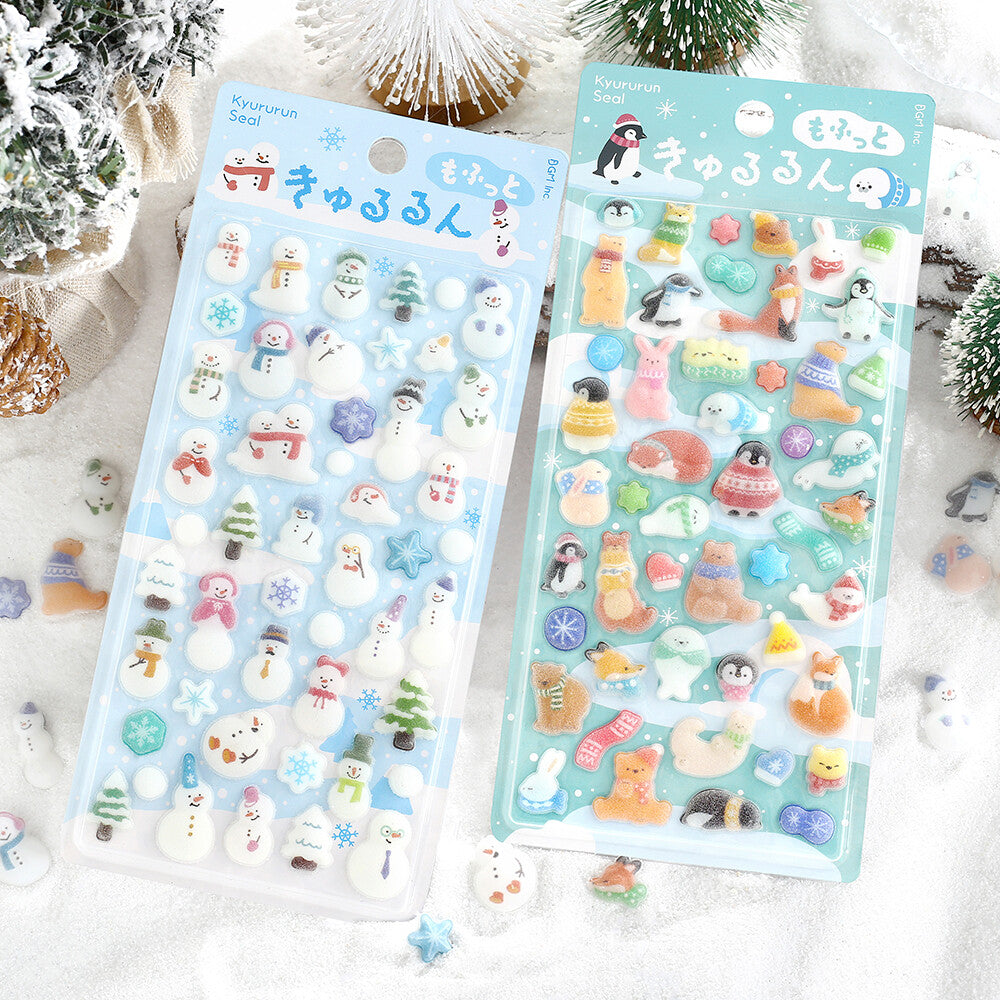 Two packages of cute winter-themed stickers displayed on a snowy background with miniature pine trees and ornaments. The left package contains snowman-themed stickers, and the right package contains penguin and fox and other animal-themed stickers.