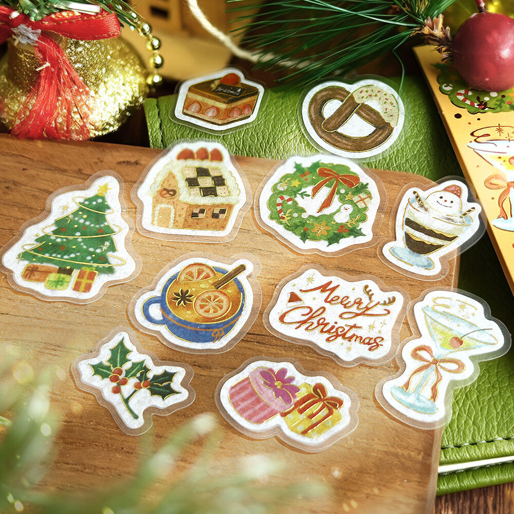 A set of Christmas-themed die-cut stickers displayed on a wooden surface with festive holiday decorations in the background. The stickers feature illustrations of a Christmas tree, gingerbread house, pretzel, wreath, hot chocolate, martini glass, gift boxes, holly, and a 'Merry Christmas' message.