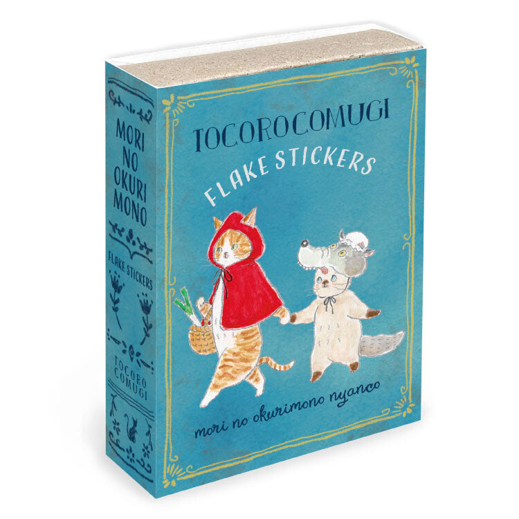 A boxed set of TocoroComugi Flake Stickers, designed to look like a vintage book with a teal-blue cover and spine, featuring illustrations of a cat in a red hood and a white dog.