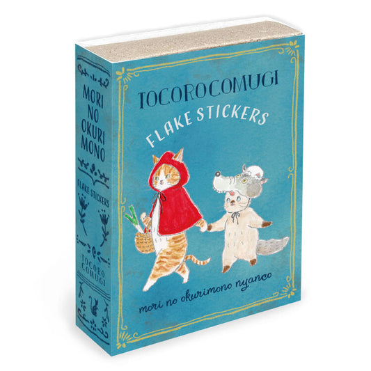 A boxed set of TocoroComugi Flake Stickers, designed to look like a vintage book with a teal-blue cover and spine, featuring illustrations of a cat in a red hood and a white dog.