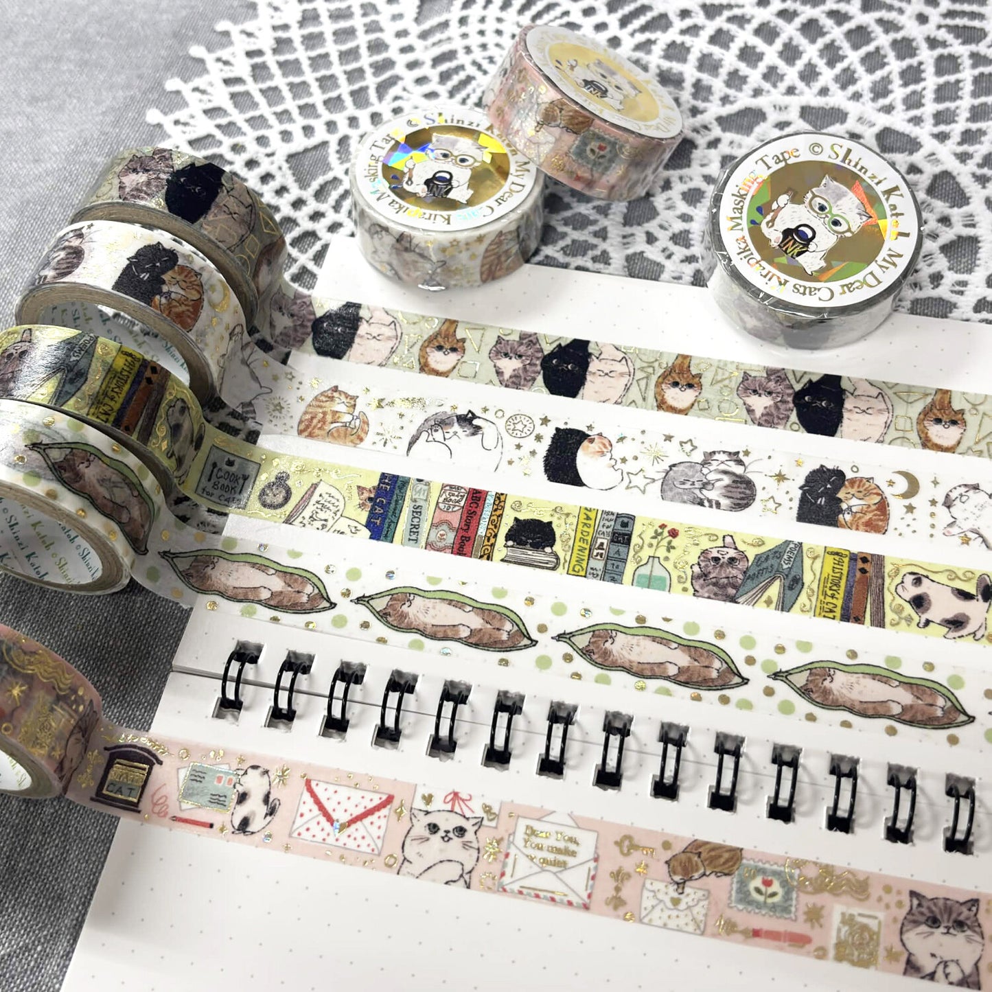 A styled flat-lay of decorative washi tape rolls and unrolled strips featuring illustrated cats, books, envelopes, and small whimsical motifs, arranged on a white notebook set on a white lace doily.