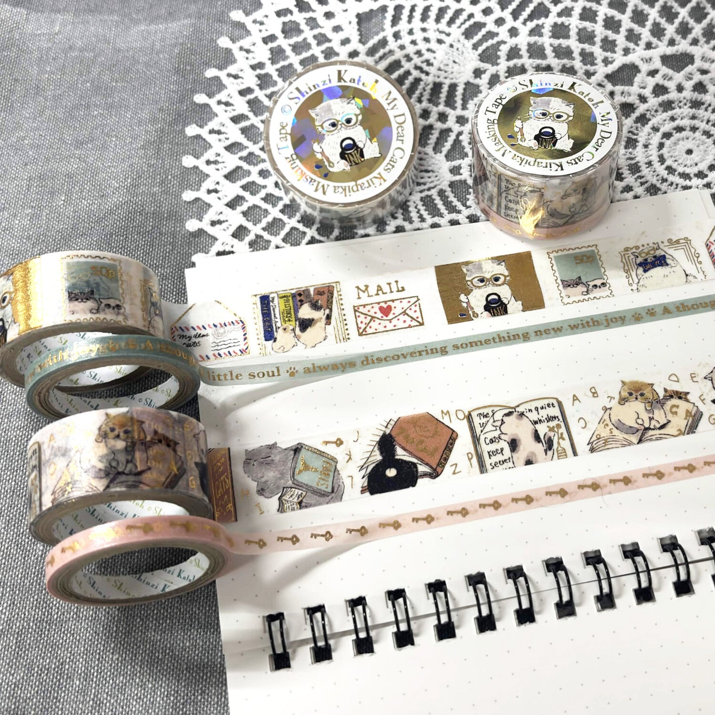 Washi tape rolls with vintage-style cat illustrations and postage stamp motifs, displayed on a white dotted notebook over a gray textured fabric with a white lace doily.