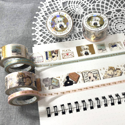 Washi tape rolls with vintage-style cat illustrations and postage stamp motifs, displayed on a white dotted notebook over a gray textured fabric with a white lace doily.