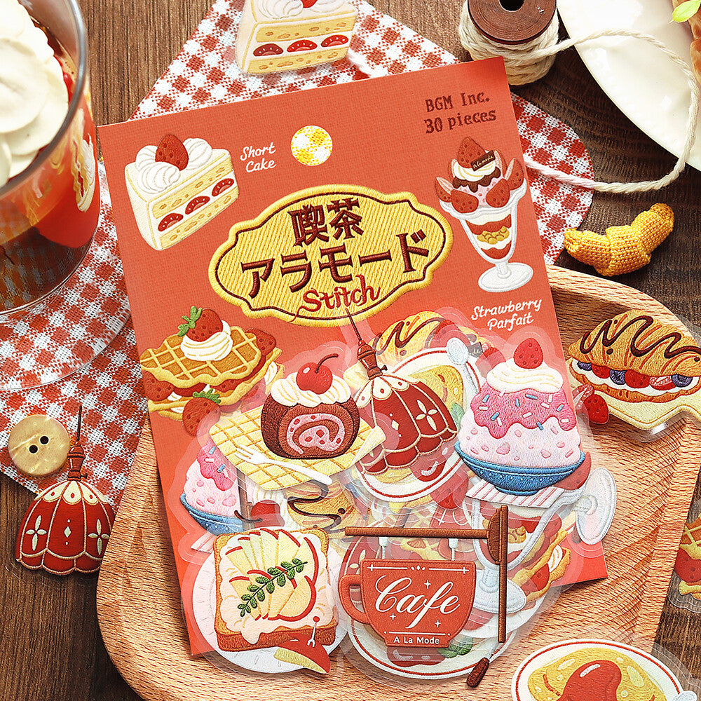 A sheet of strawberry-themed cake and dessert stickers from BGM Inc. is displayed on a wooden tray alongside a matching red-and-white checkered napkin, a glass of iced coffee, and other cake illustrations.