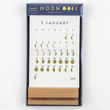 A boxed January 2027 calendar featuring a white page with a moon phase grid and dates, in a dark navy package with gold moon illustrations and a small wooden stand visible at the bottom.