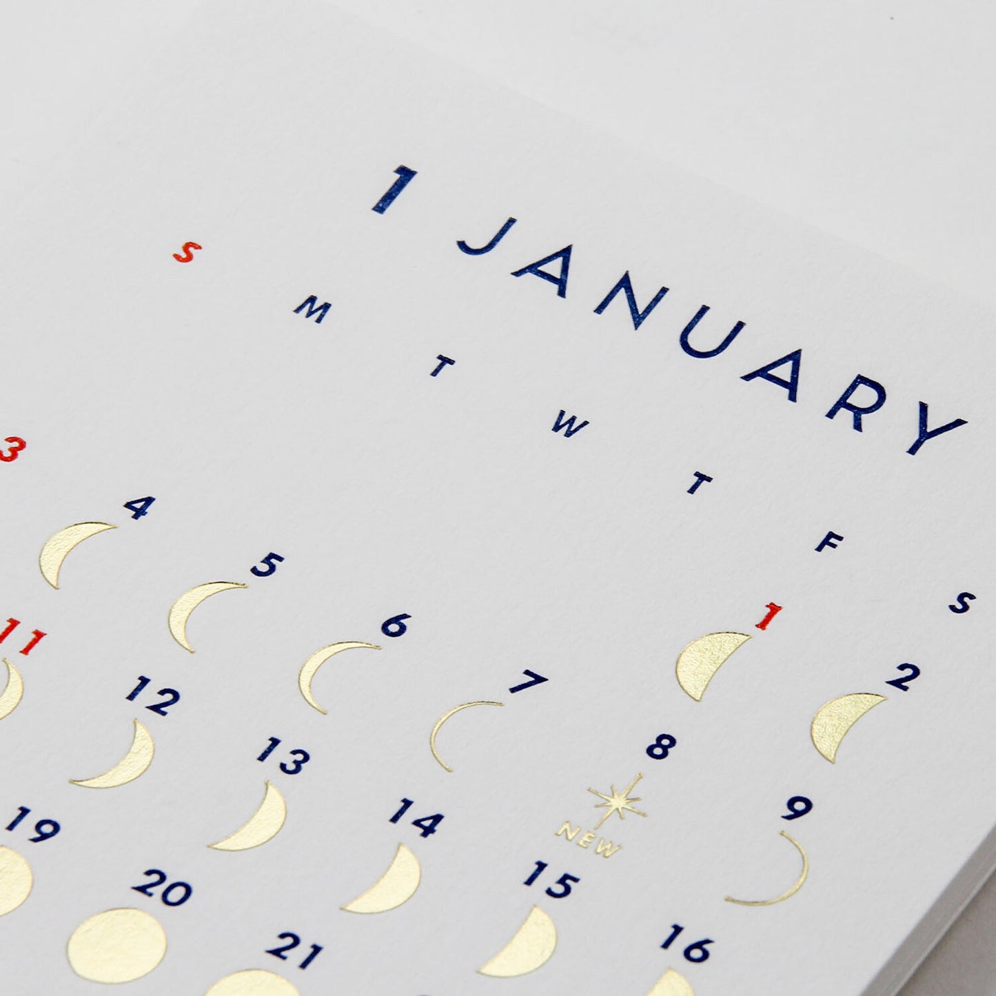 A close-up of a white calendar page showing January with gold foil moon phases, blue weekday letters, and a red '1' for the first day.