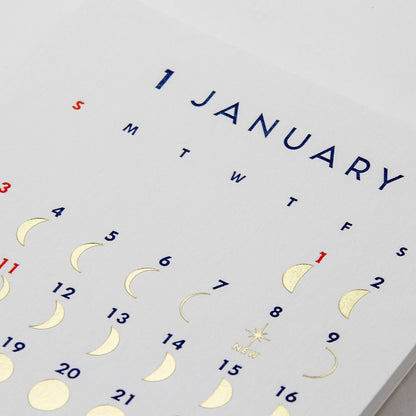A close-up of a white calendar page showing January with gold foil moon phases, blue weekday letters, and a red '1' for the first day.