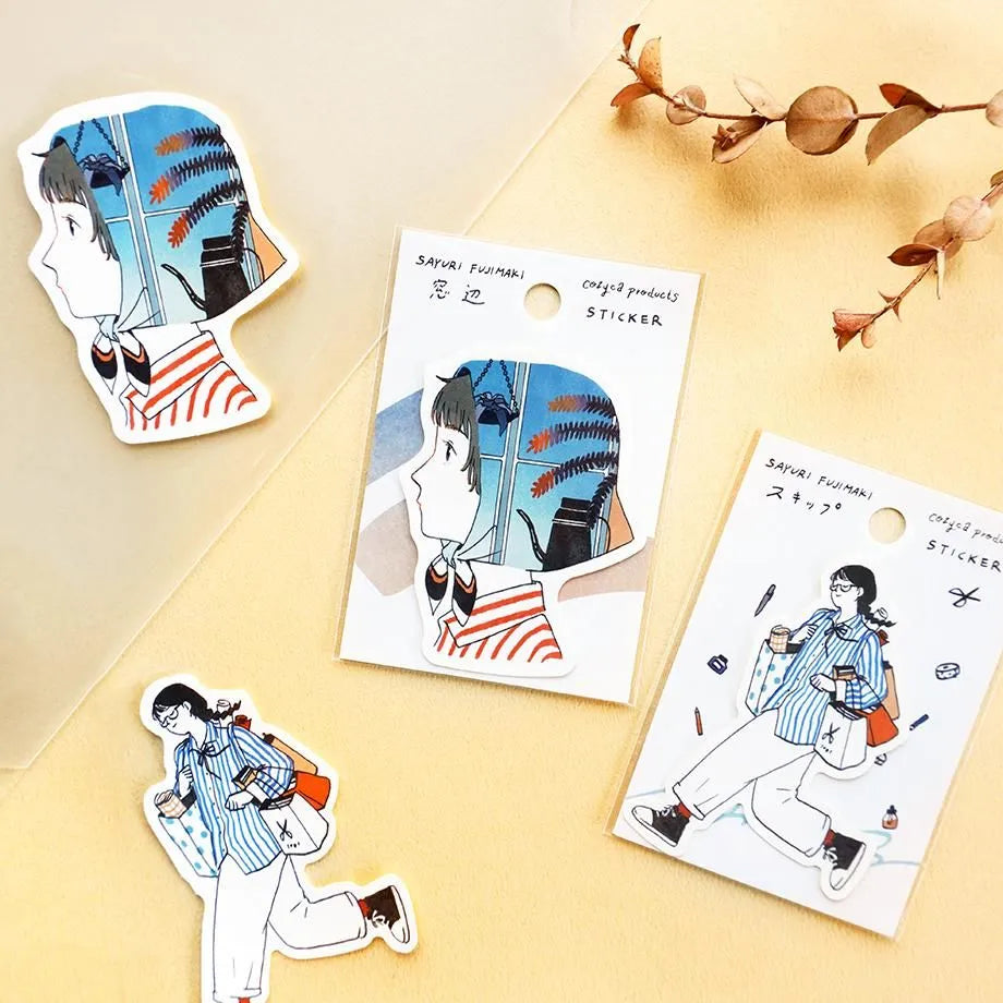 Three illustrated die-cut stickers displayed on a warm beige background, including two printed sticker cards and one loose character sticker. The artwork shows a girl in a blue-and-white striped shirt with red and orange accents in a stationery-themed scene.