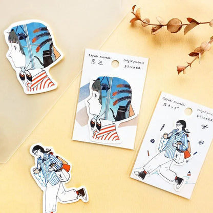 Three illustrated die-cut stickers displayed on a warm beige background, including two printed sticker cards and one loose character sticker. The artwork shows a girl in a blue-and-white striped shirt with red and orange accents in a stationery-themed scene.
