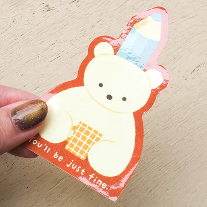 A hand with glittery brown nail polish holds a die-cut sticker featuring a cute cream-colored bear wearing a blue and yellow striped party hat and holding a yellow checkered mug. The sticker has a red-orange border and the text 'you'll be just fine.