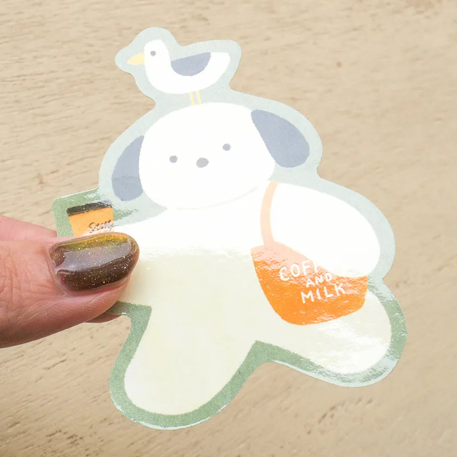 A die-cut sticker featuring a cute cartoon dog character with a bird on its head, being held by a hand with glittery brown nail polish. The sticker has a white body, gray ears, a light green outline, and an orange bag that reads 'COFFEE AND MILK'.