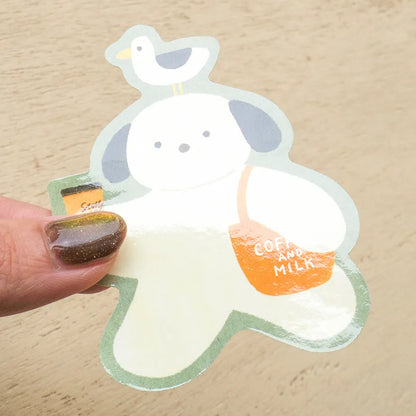 A die-cut sticker featuring a cute cartoon dog character with a bird on its head, being held by a hand with glittery brown nail polish. The sticker has a white body, gray ears, a light green outline, and an orange bag that reads 'COFFEE AND MILK'.