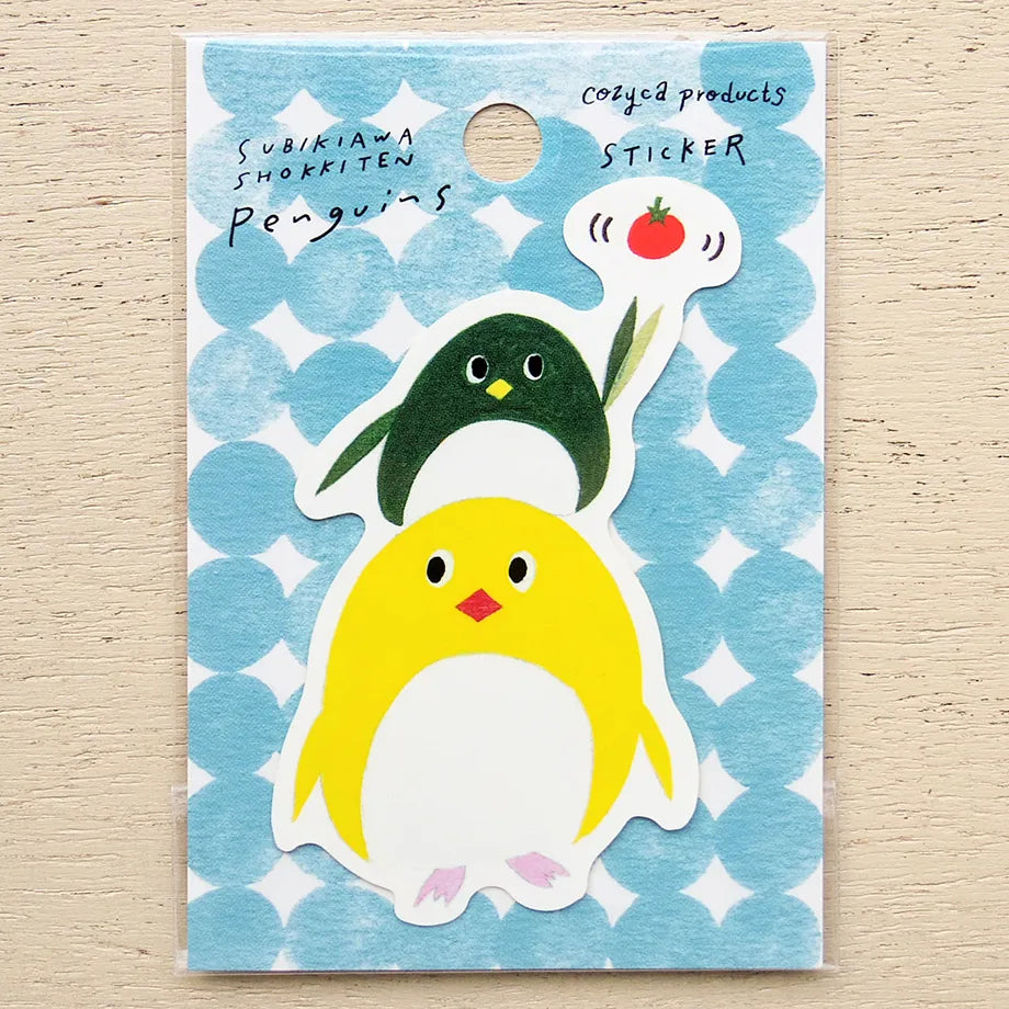 A sticker set featuring two cute cartoon penguins on a blue patterned backing card. The front sticker shows a yellow penguin with pink feet and a red beak, while the back sticker shows a dark green penguin with a speech bubble containing a red tomato.