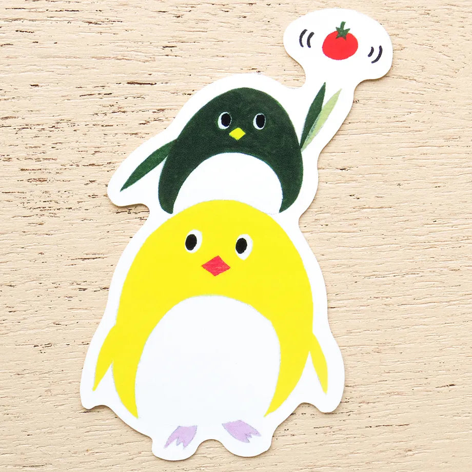 A die-cut sticker featuring two cute cartoon penguins on a light wood background. The foreground penguin is bright yellow with a white belly, black eyes, a small red beak, and light purple feet.