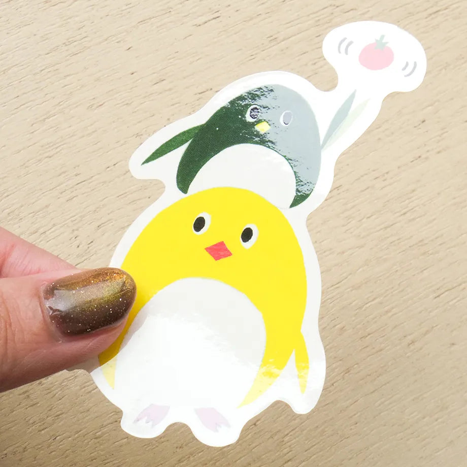A die-cut sticker of a cute cartoon penguin being held by a hand with glittery brown nail polish, set against a light wood grain background.