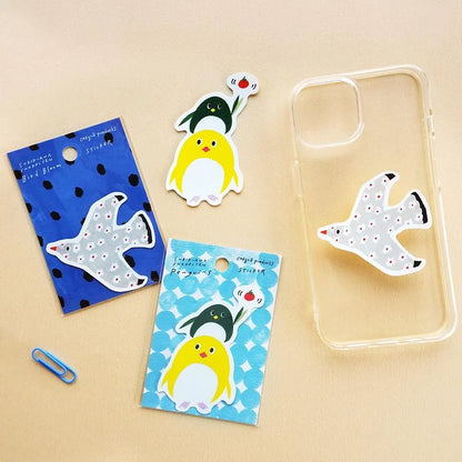 Three cute cartoon penguin-themed phone accessories are arranged on a beige background: a clear phone case with a white penguin design, a blue patterned backing card with a white penguin sticker, a yellow-and-white penguin sticker, and a blue-and-black penguin sticker on a light blue patterned backing card. A small blue paperclip is also present as a prop.