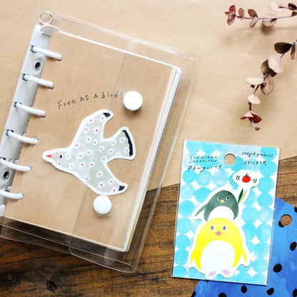 A clear plastic ring-bound notebook with a kraft paper cover and a bird-shaped sticker, shown on a wooden surface next to a blue card with colorful penguin stickers.