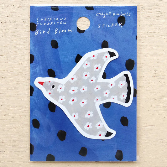 A bird-shaped sticker on a blue backing card with black polka dots and white handwritten text reading 'Subkiawa Shokiten Bird Bloom' and 'Cozyca products Sticker'. The sticker is light gray with white floral patterns and small red and black accents.