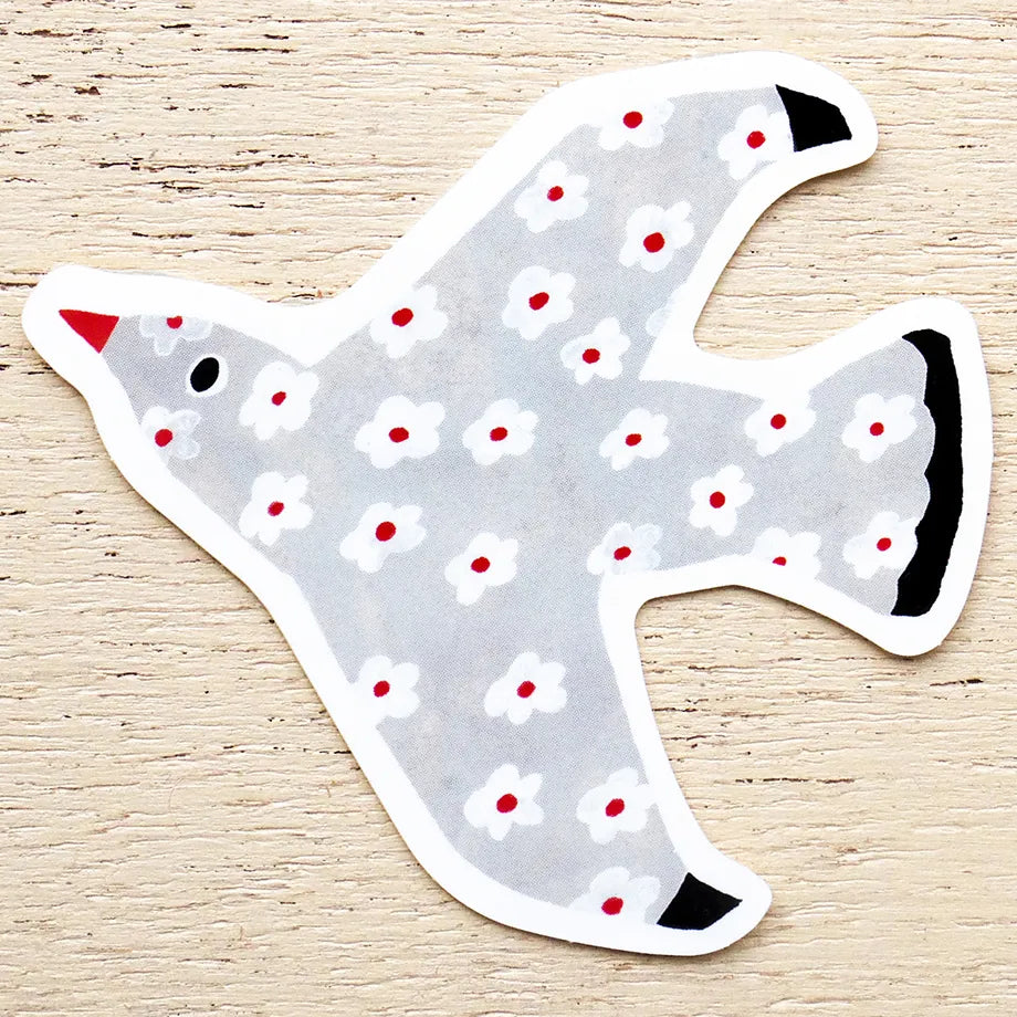 A die-cut sticker shaped like a dove, featuring a light gray body with a white and red floral pattern, a red beak, and black accents, displayed on a light wood background.