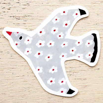 A die-cut sticker shaped like a dove, featuring a light gray body with a white and red floral pattern, a red beak, and black accents, displayed on a light wood background.