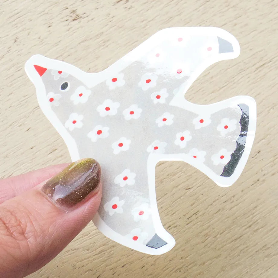 A die-cut sticker shaped like a bird, held by a hand with glittery nail polish against a light wood background. The sticker is white with small white flowers, red dots, and a red beak.