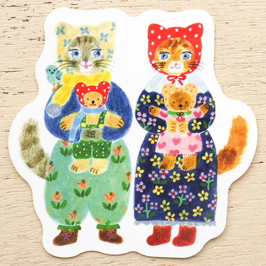 A die-cut sticker featuring a whimsical illustration of a gray cat in a blue outfit holding a teddy bear, next to an orange tabby cat in a red polka-dot bonnet and dark floral dress holding a teddy bear, shown on a light wood background.