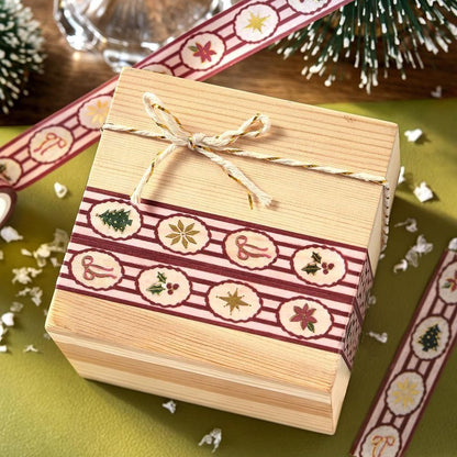 A square wooden gift box with a light natural wood finish, decorated with a red and white holiday patterned washi tape and set on a green surface with festive props.