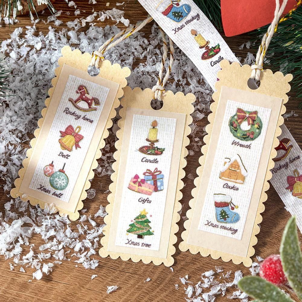 Three scalloped-edge Christmas-themed bookmark tags laid on a wooden surface with white shavings and holiday greenery. Each tag has a cream center masking tape panel with small illustrated holiday motifs and words like rocking horse, bells, ornament, candle, gift, Christmas tree, wreath, cookie, and Santa stocking.