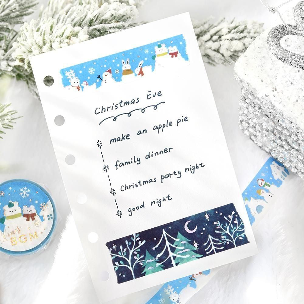 A white planner insert sheet with handwritten 'Christmas Eve' text and a festive winter-themed decorative washi tape strip at the bottom, styled on a white surface with snowy greenery, a silver ornament, and a round pin badge.