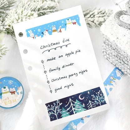 A white planner insert sheet with handwritten 'Christmas Eve' text and a festive winter-themed decorative washi tape strip at the bottom, styled on a white surface with snowy greenery, a silver ornament, and a round pin badge.